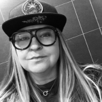 Selfie of a woman wearing a baseball cap with a circular emblem, oversized glasses, and long hair (black and white). Necklaces and a dark top complete the look outside a tiled wall backdrop.
